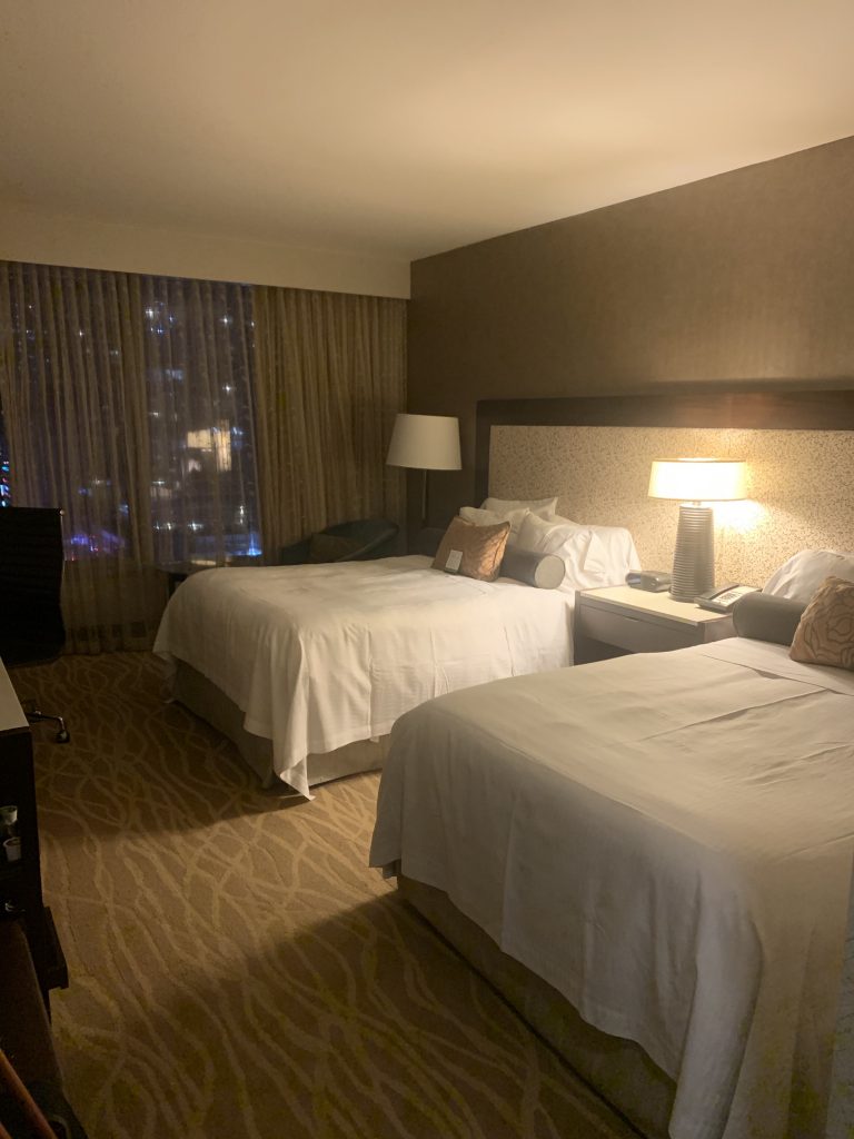 Photo of our hotel room. Two double beds against a lovely brown yet sophisticated wallpaper with a lamp and table between the two beds. floor to ceiling windows just beyond both beds overlooking new york city.