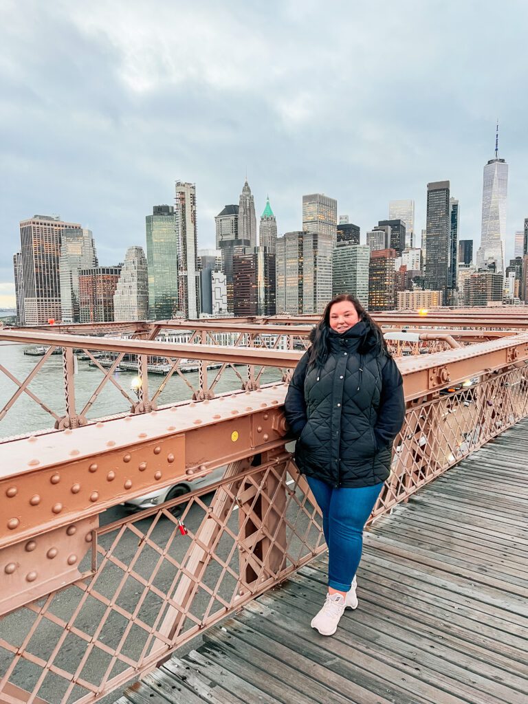 photo of Han on the brooklyn bridge in New york, wearing a black jacket with blue jeans and white trainers with the new york city skyline in the background. The sky is dusk and cloudy.