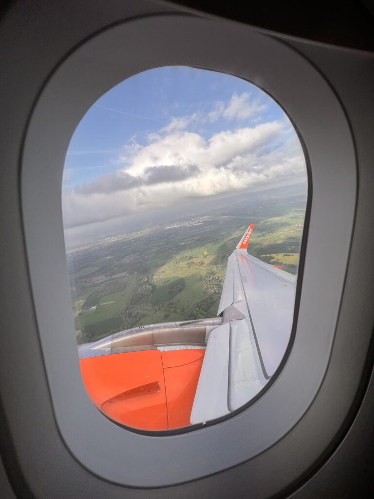 photo of a plane window with the wing of the plane in the middle of the window and the greenery of down below behind the wing of the plane. the plane is an easyjet plane so the wing is white with the tip being bright orange
