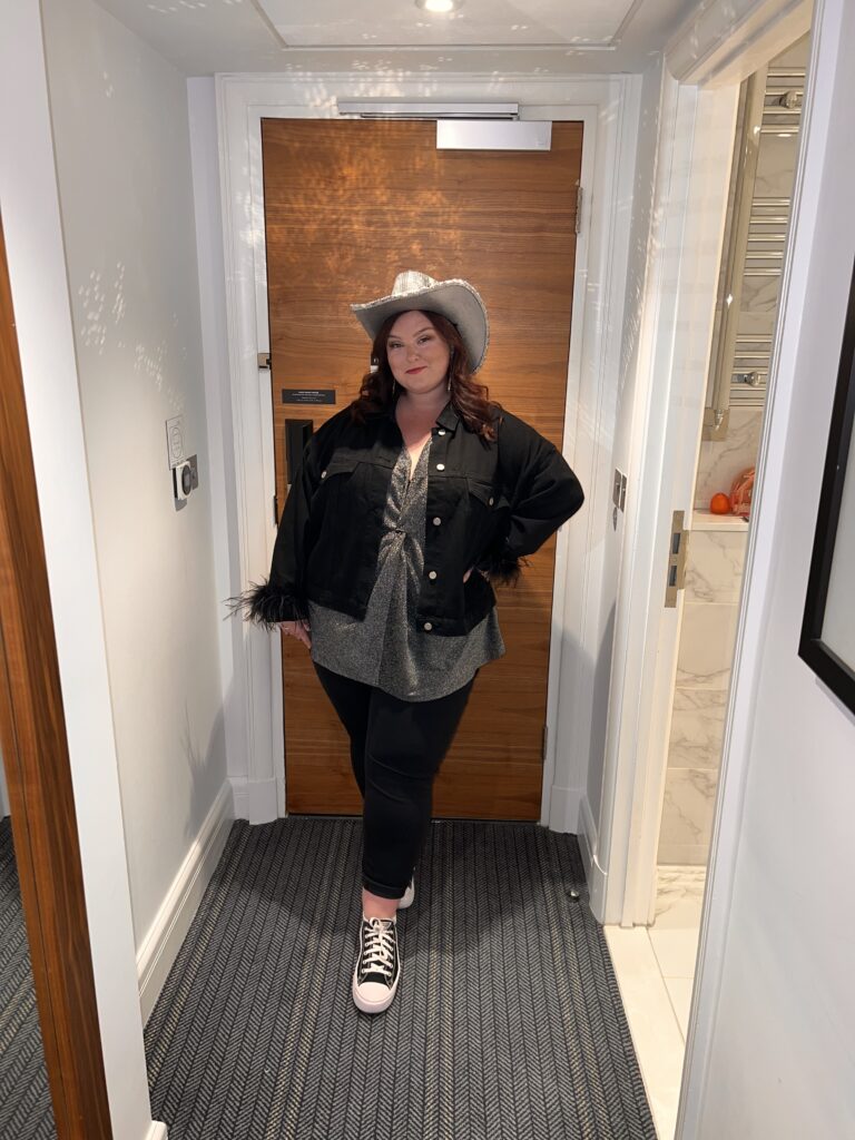 photo of hannah stood infront of a door, wearing a silver cowboy hat, a silver top and black jeans