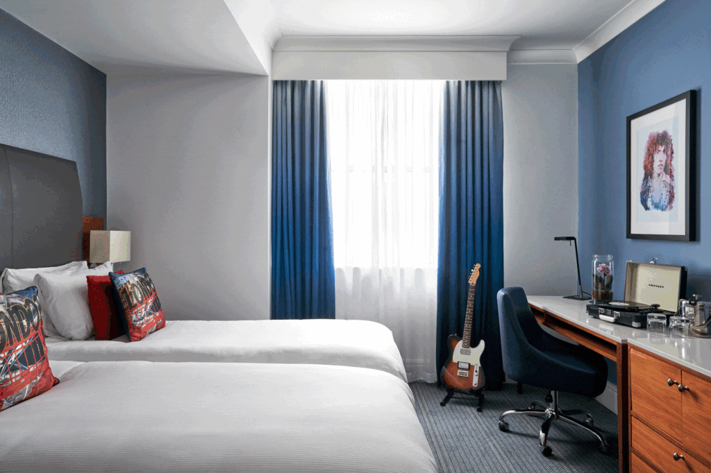photo of the hotel room. There are two twin beds in the room, with a window with blue curtains and a desk at the end of the beds. The walls are a light navy colour with one wall being white with red, white and blue cushions on the bed