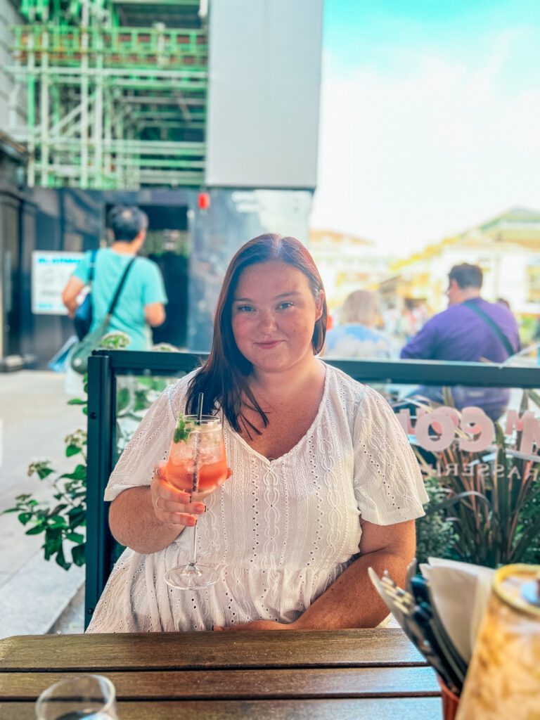 this photo is of hannah sat across the table outside in a restaurant in central london, wearing a white peplum top holding a glass of wine