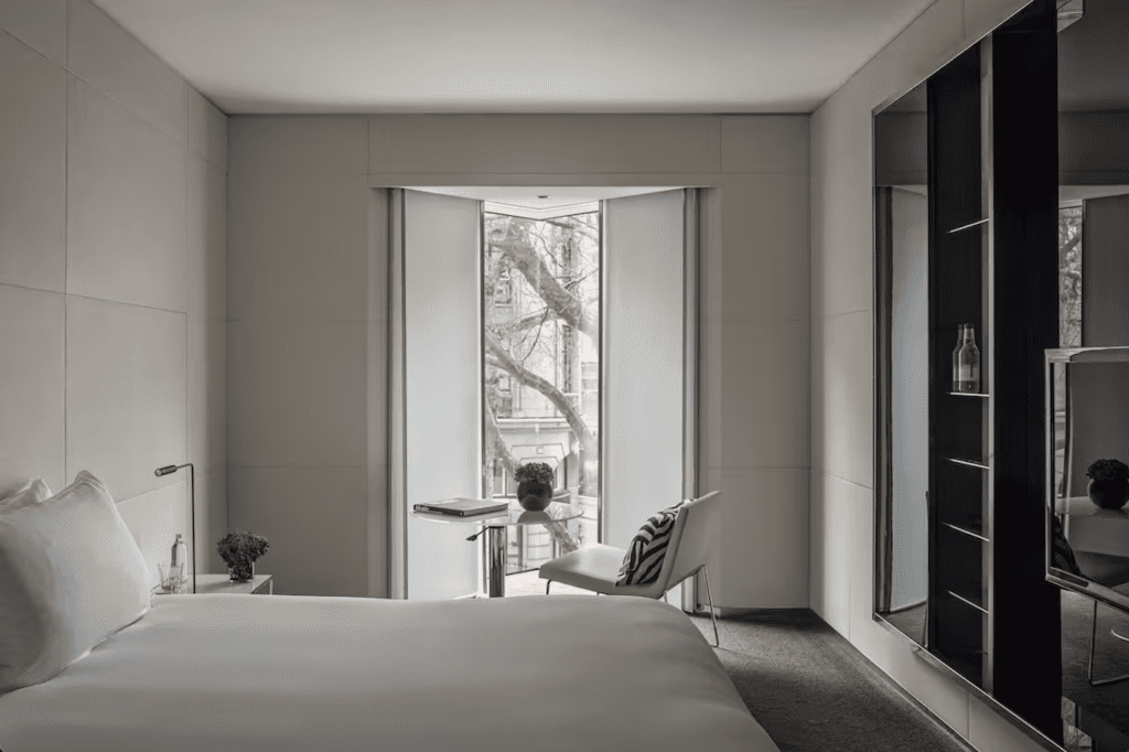 this photo shows a white room with a white kingsize bed in the centre with a large black tv opposite the end of the bed with a table and chairs infront of the floor to ceiling windows