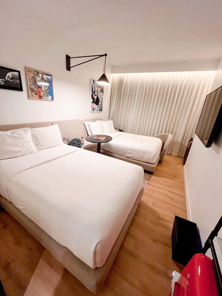 photo of the two double beds within the virgin hotel room. The walls are white with cream floor to ceiling curtains closed across the window with art on the walls and the headboard is a neutral colour with white bedding