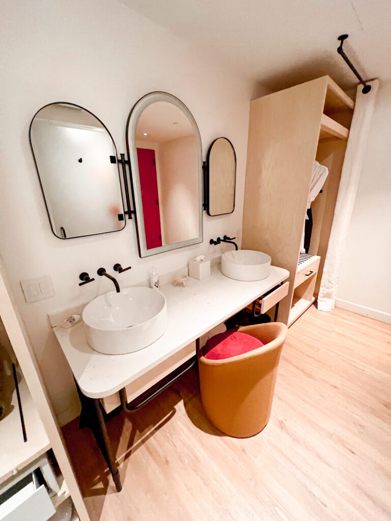photo of the bathroom vanity area within the virgin hotel room. it has a white countertop with two white sinks with black taps as well as a mirror on the wall with two smaller mirrors either side of the main mirror