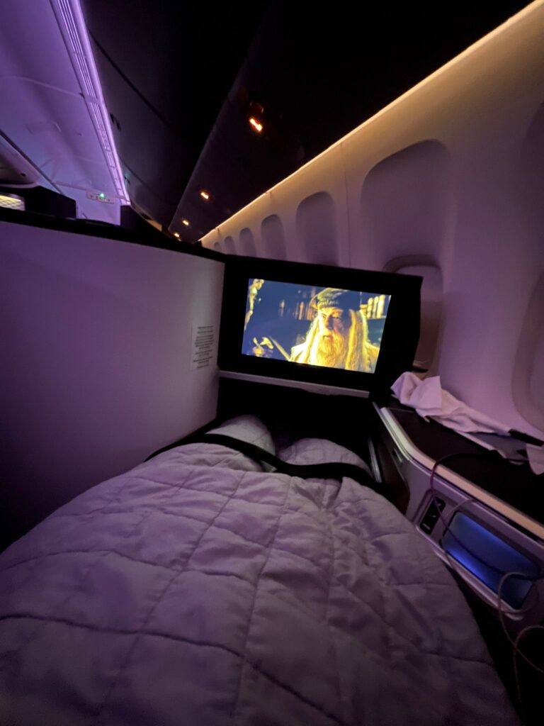 business class seat lay flat wioth the blanket over hannah and the TV is playing a movie