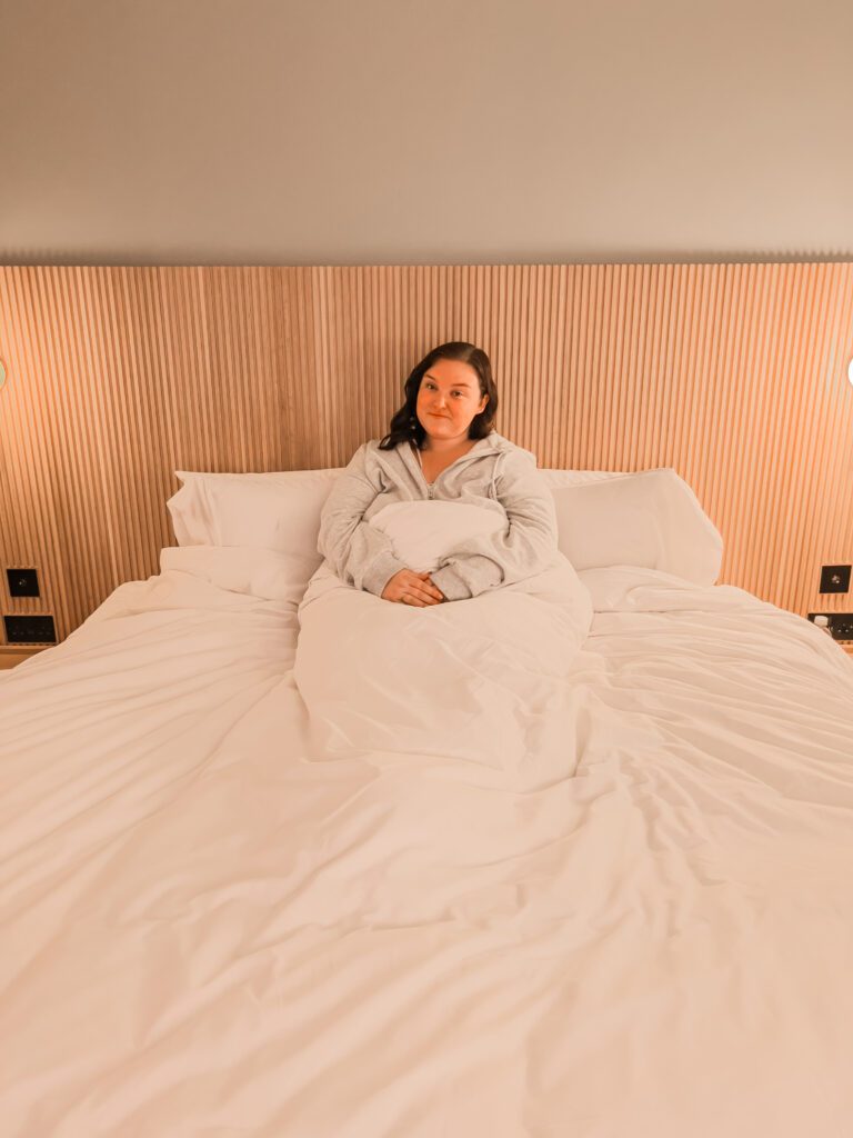 hannah sat on the bed in mollies motel. there are wooden panels behind her and the bedding is white